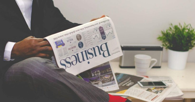 man reading newspaper while sitting near table with smartphone and cup