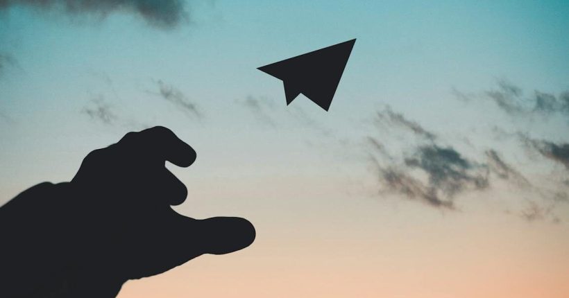A silhouette of a hand launching a paper plane against a colorful dusk sky with a crescent moon.