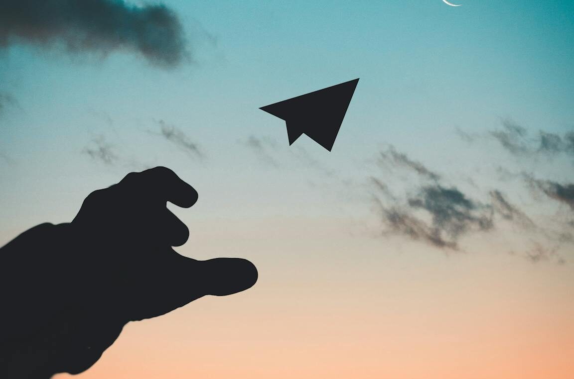 A silhouette of a hand launching a paper plane against a colorful dusk sky with a crescent moon.