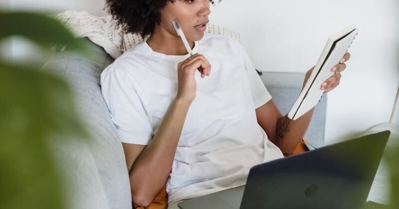 Focused young woman studying at home with a laptop and notepad, sitting comfortably on a couch.