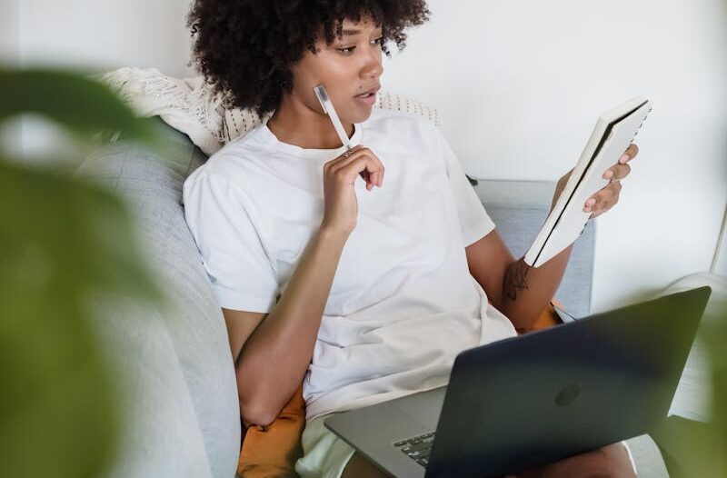 Focused young woman studying at home with a laptop and notepad, sitting comfortably on a couch.