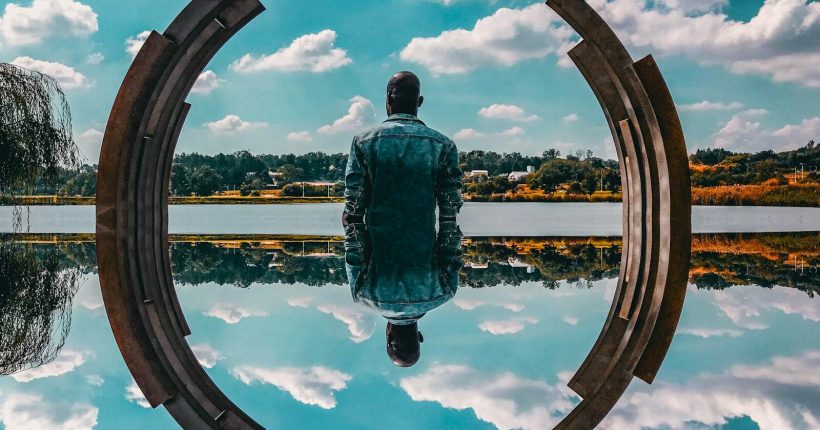 A man in a denim jacket sits by abstract sculpture reflecting on the water under a blue sky.