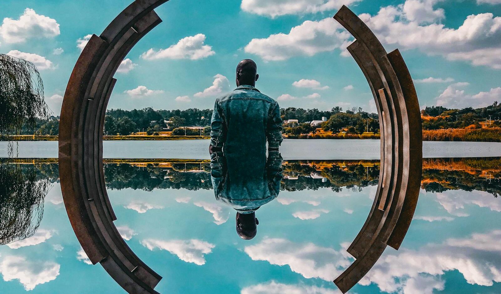 A man in a denim jacket sits by abstract sculpture reflecting on the water under a blue sky.