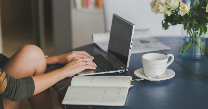 Female freelancer using laptop with coffee at home office desk, surrounded by roses and a planner.