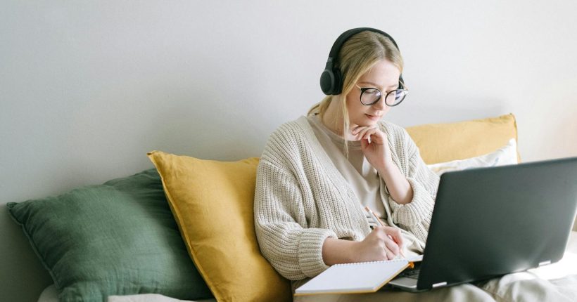 A focused woman in glasses and headphones works on a laptop from a cozy bed.