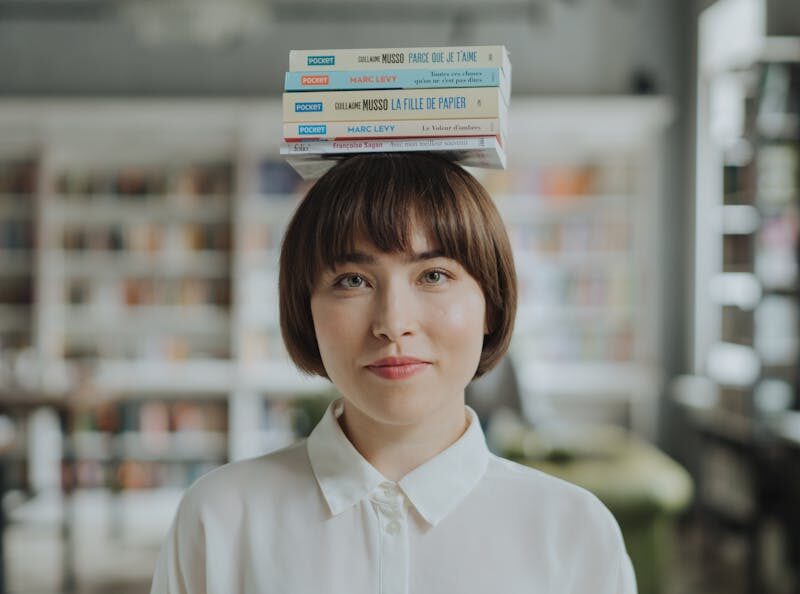 Young woman in a library balancing books on her head, wearing a white shirt.