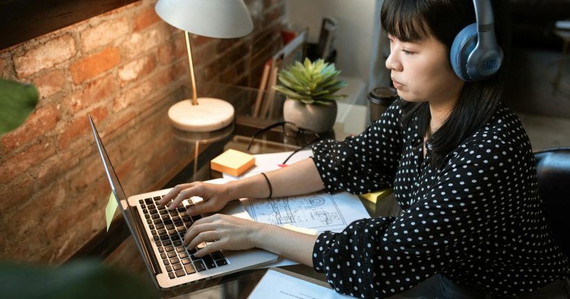 Focused woman wearing headphones, working on a laptop in a cozy home office setup with natural light.