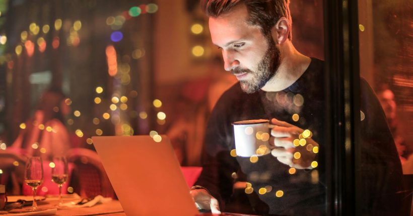 A young man with a beard is working on a laptop at a cozy cafe with warm lighting, holding a coffee cup.