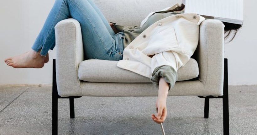 Tired woman in casual attire falls asleep on sofa with a notebook on face and pen in hand.