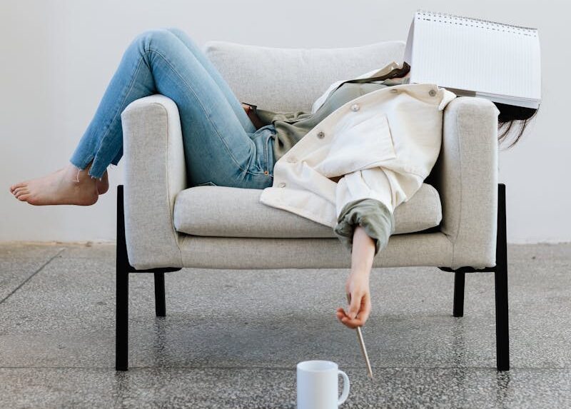 Tired woman in casual attire falls asleep on sofa with a notebook on face and pen in hand.