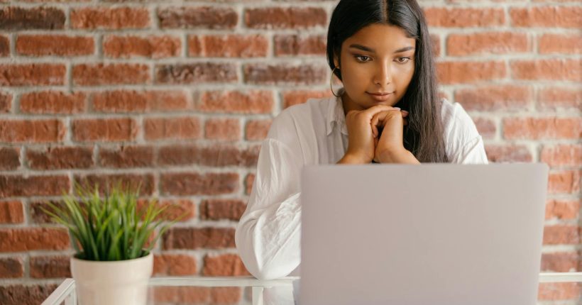 Young woman deeply focused on her laptop studying indoors against a brick wall backdrop.