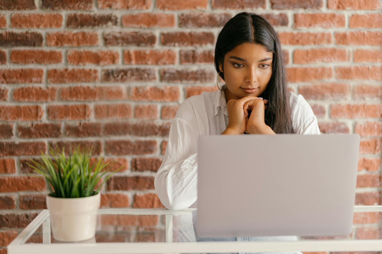 Young woman deeply focused on her laptop studying indoors against a brick wall backdrop.