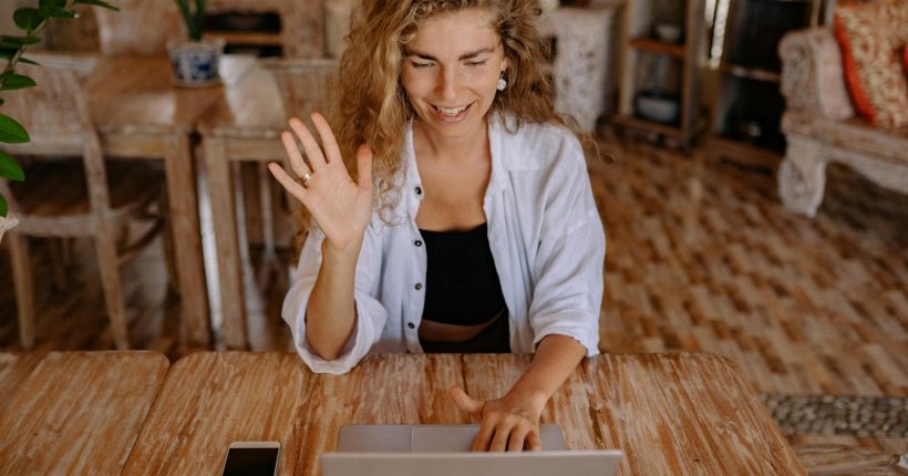 Smiling woman waves during a video call on her laptop in a comfortable indoor setting.