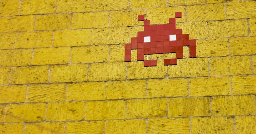 Street art featuring a retro pixelated space invader on a vivid yellow brick wall.
