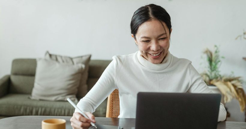A cheerful woman uses a laptop and tablet for a video call, working remotely in a cozy living room.
