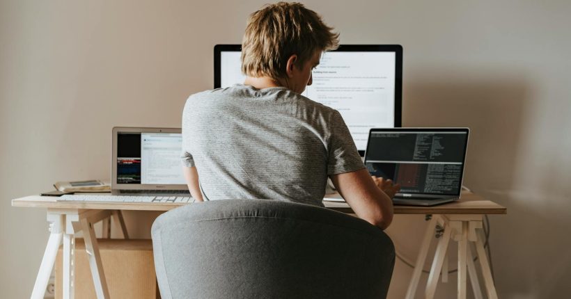 Young man coding at home office with multiple laptops, representing remote work and technology.