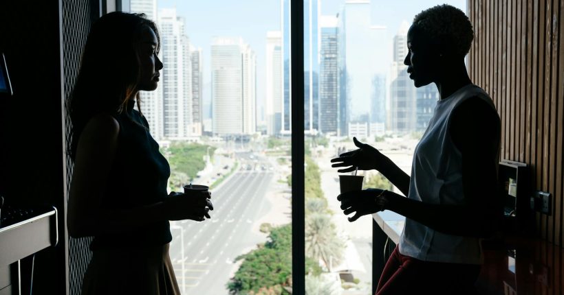 Two businesswomen engage in discussion by a large window overlooking a cityscape. A corporate setting.