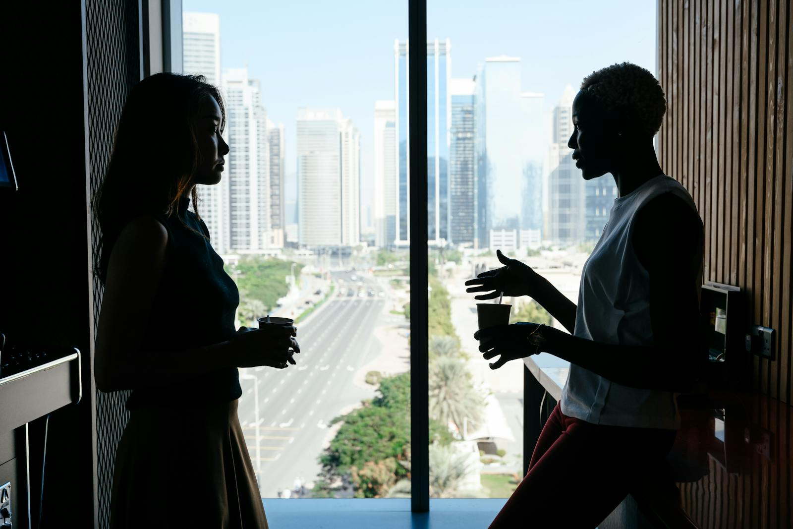 Two businesswomen engage in discussion by a large window overlooking a cityscape. A corporate setting.
