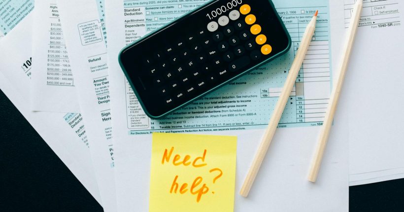 Top view of financial papers with a calculator, pencils, and a note saying 'Need help?' indicating tax or accounting assistance.