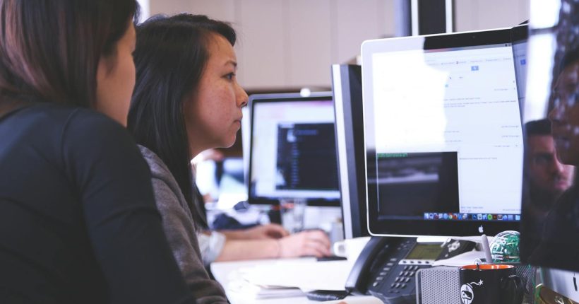 Two women working together on code at a computer in a modern office setting.