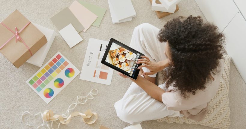 Woman using tablet surrounded by design tools and gift box, showcasing creativity and online work.