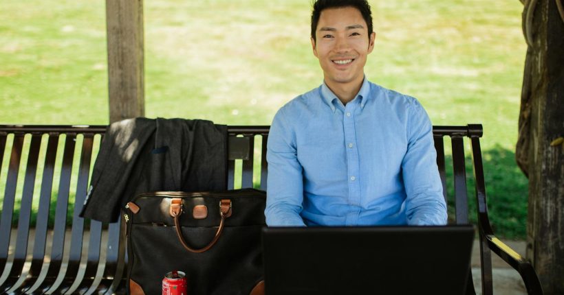 Asian man in casual attire working on laptop outside on a sunny day, enjoying remote work lifestyle.