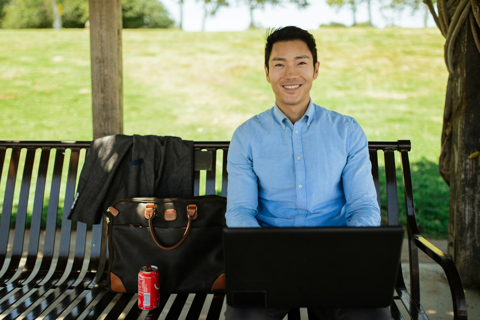 Asian man in casual attire working on laptop outside on a sunny day, enjoying remote work lifestyle.