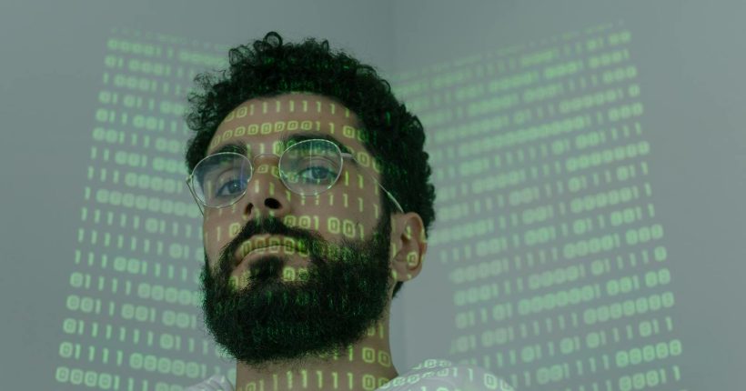 A man with a beard and eyeglasses looking at the camera, illuminated by binary code projection.