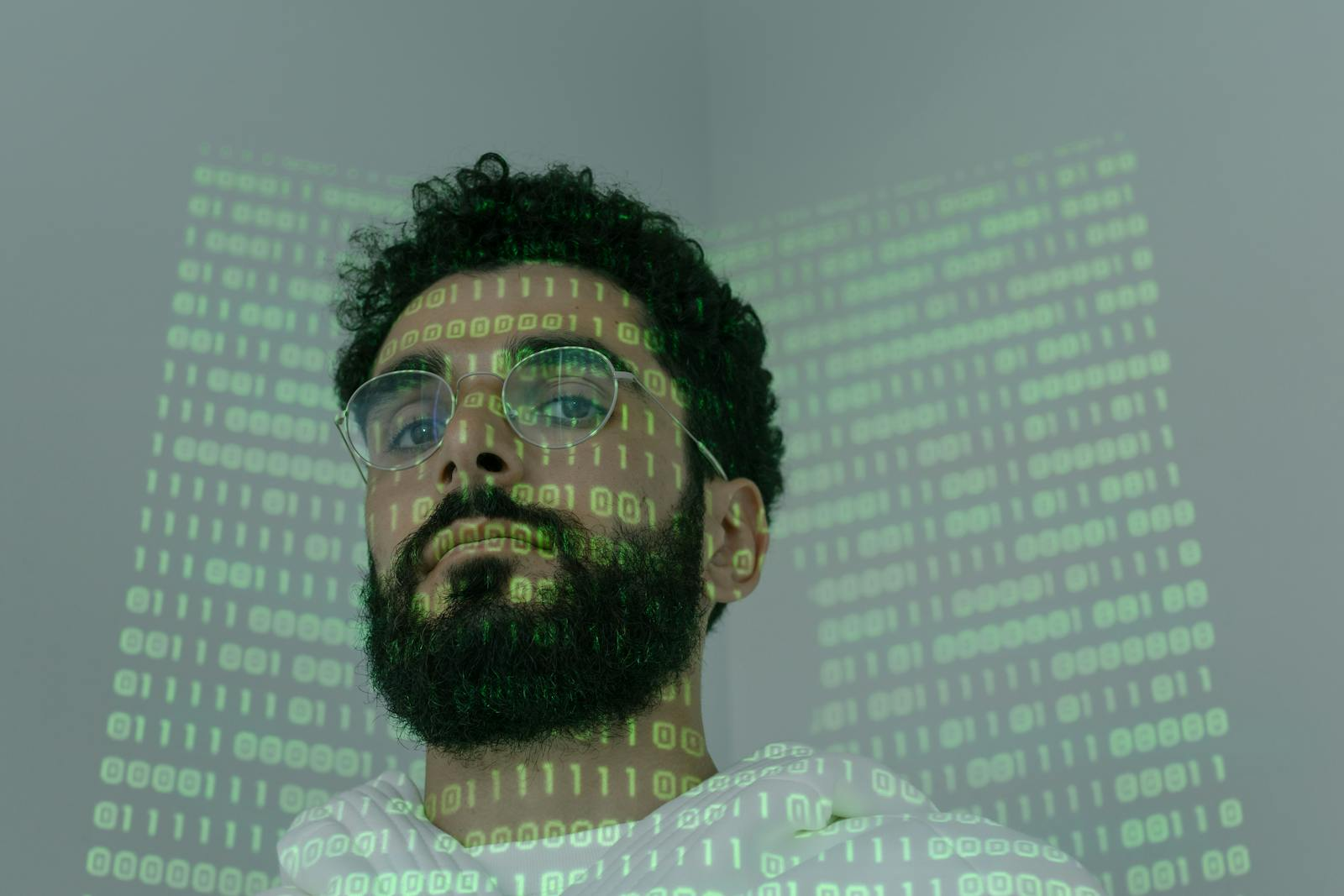 A man with a beard and eyeglasses looking at the camera, illuminated by binary code projection.