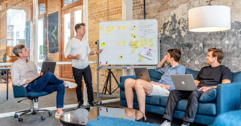 three men sitting while using laptops and watching man beside whiteboard