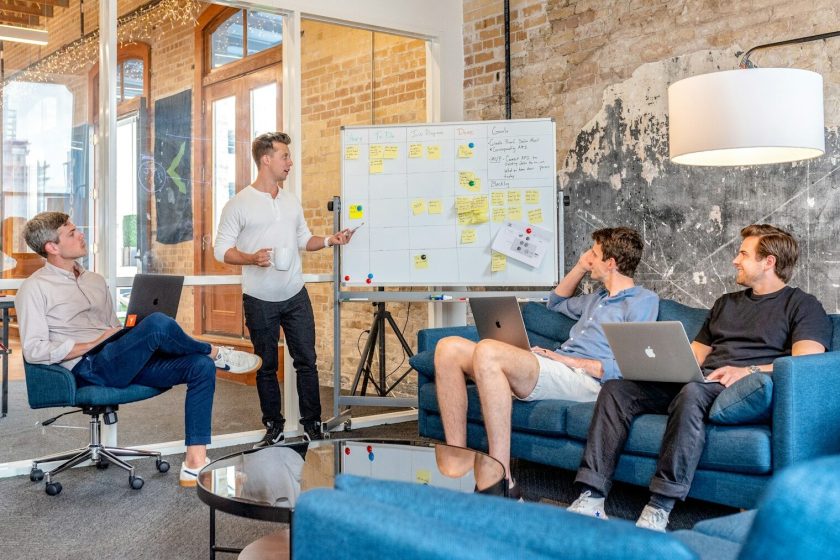 three men sitting while using laptops and watching man beside whiteboard