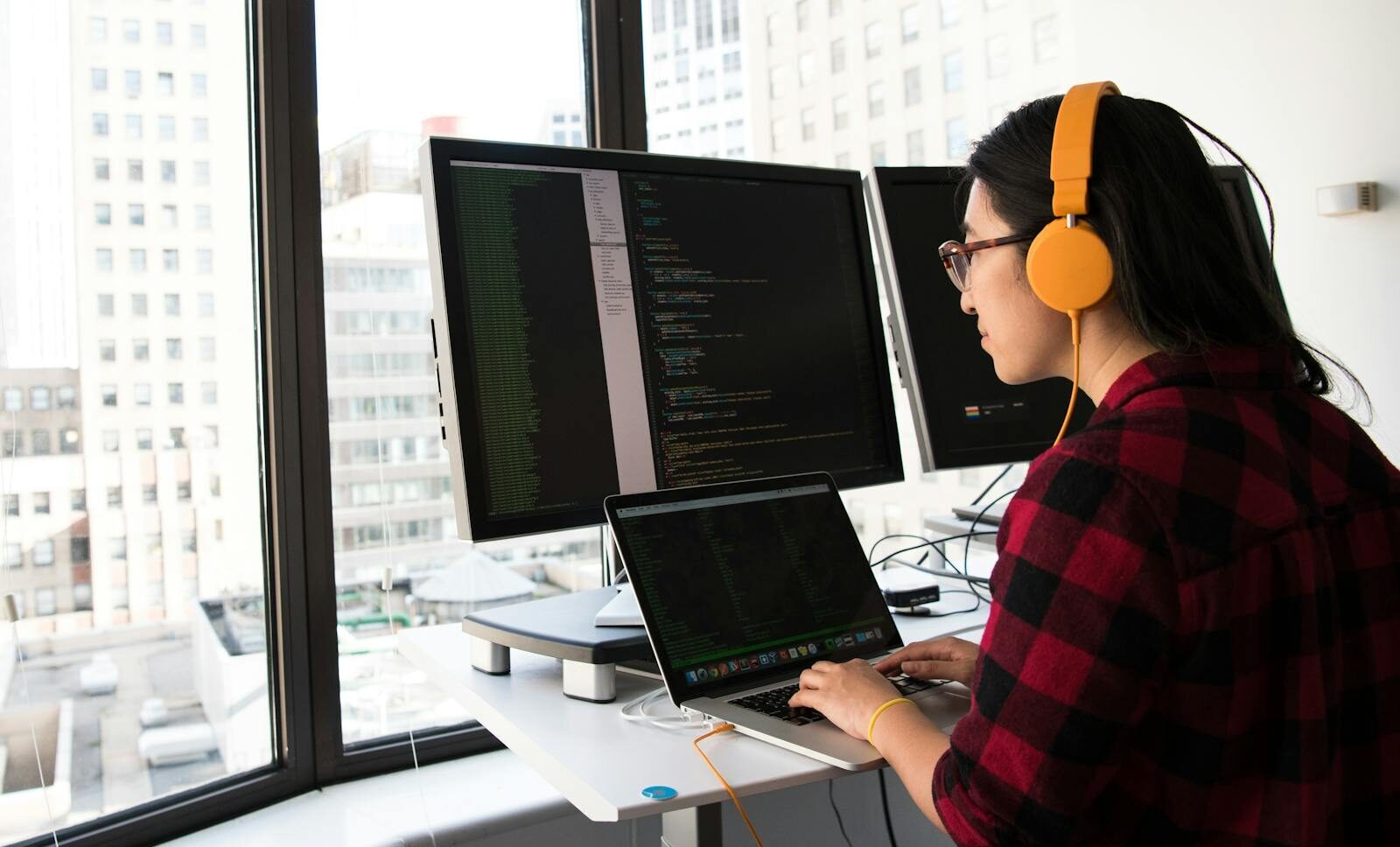 Woman programming on a laptop at a standing desk in an office with large windows.