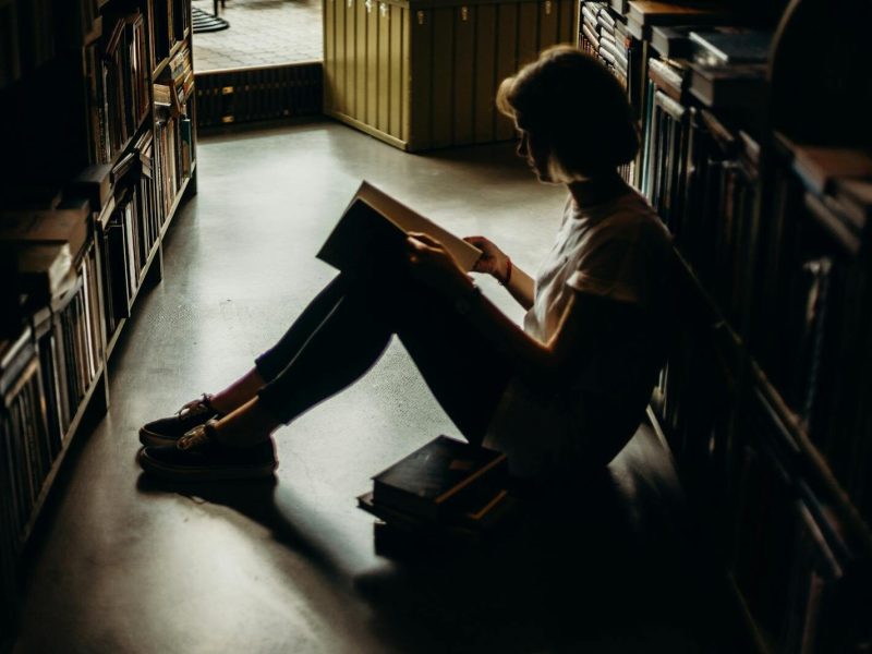 A young woman engrossed in a book, sitting on the floor of a quiet library, creating a cozy reading atmosphere.