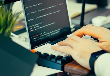 Close-up view of a developer typing code on a keyboard with a computer screen showing scripts.