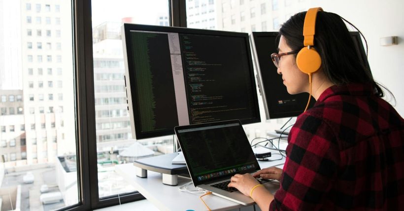 Woman programming on a laptop at a standing desk in an office with large windows.