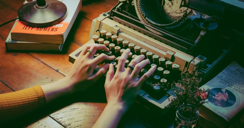 A cozy scene with a vintage typewriter and hands typing indoors beside books and flowers.