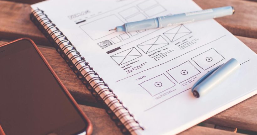 Close-up of a notebook with wireframe sketches and a smartphone on a wooden desk.