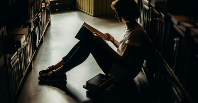 A young woman engrossed in a book, sitting on the floor of a quiet library, creating a cozy reading atmosphere.