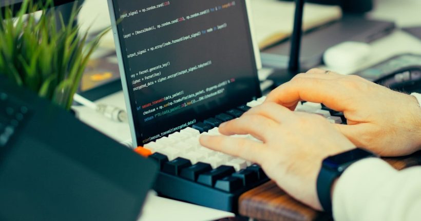 Close-up view of a developer typing code on a keyboard with a computer screen showing scripts.