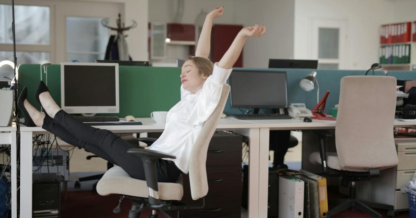Side view of cheerful female employee in formal outfit sitting on office chair with crossed legs on desk and stretching while resting during work with closed eyes