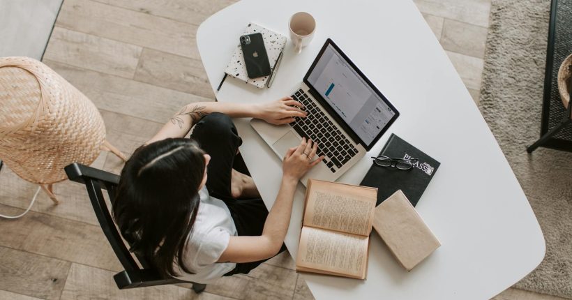 Overhead view of a woman using a laptop at a home desk, surrounded by books, a phone, and a cup.
