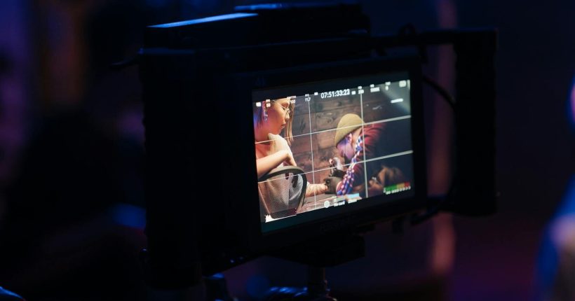Film crew adjusting camera settings on set highlighted by neon lights.