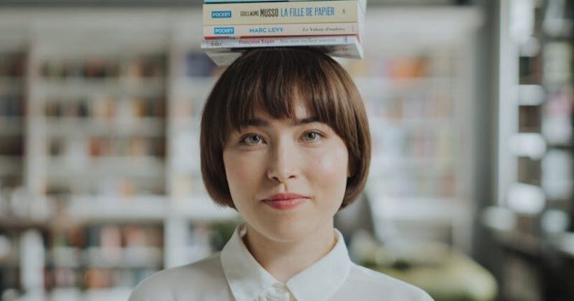 Young woman in a library balancing books on her head, wearing a white shirt.