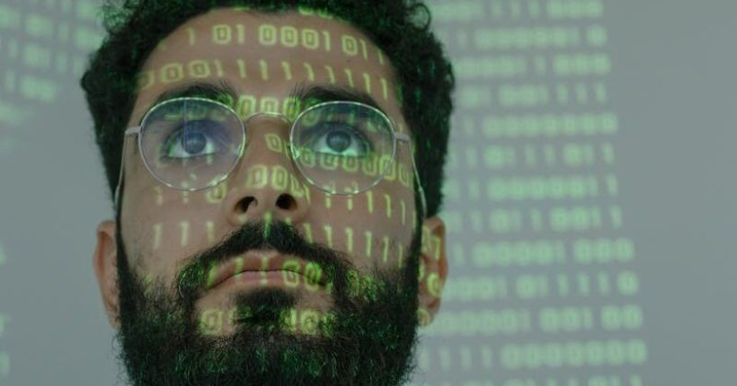 A bearded man with eyeglasses analyzing data in a digital environment with green binary code projections.