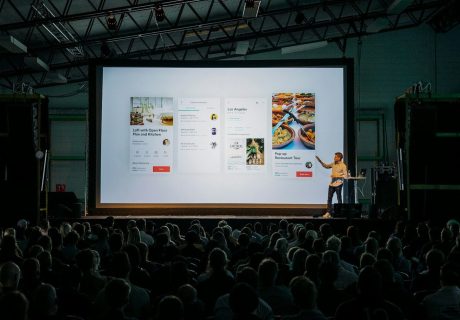person discussing while standing in front of a large screen in front of people inside dim-lighted room