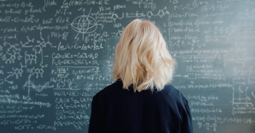 Woman with blonde hair facing a chalkboard with equations.