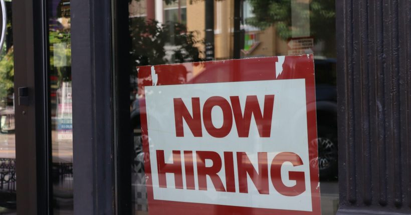 A "now hiring" sign hangs in a store window.